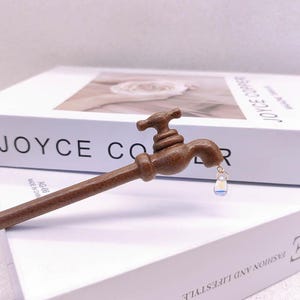 May include: A brown wooden hair stick shaped like a water tap with a small, clear, iridescent teardrop-shaped charm hanging from the spout. The tap handle is positioned at the top, and the long, slender wooden shaft extends downwards.
