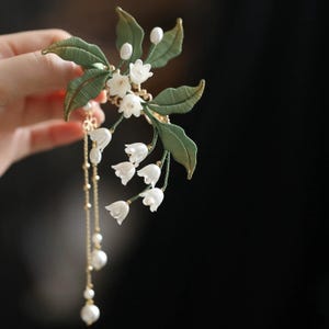 May include: A hair accessory featuring white lily of the valley flowers and textured green leaves. The piece includes delicate gold-toned chains adorned with small white beads and pearls that hang downward from the floral arrangement.
