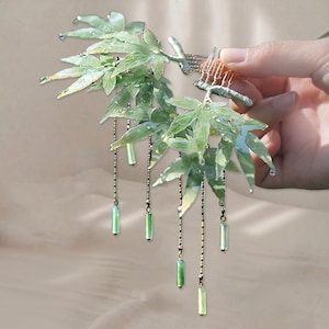 Op de afbeelding: Een haarspeld met een cluster van groene, doorschijnende esdoornbladeren, zilveren kettingen en groene kraalaccenten. De bladeren hebben een getextureerd, geaderd uiterlijk. De kam wordt door een hand vastgehouden.