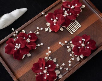 Op de afbeelding: Een verzameling bordeauxrode fluwelen bloem haaraccessoires, waaronder een kam en verschillende individuele bloemstukken. Elk stuk heeft imitatieparels en goudkleurige accenten, gerangschikt op een houten dienblad. De bloemen hebben een zachte, getextureerde uitstraling.