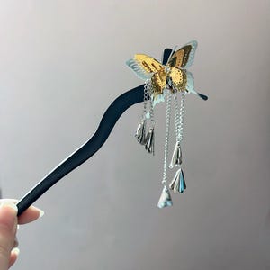 May include: A decorative hair stick featuring a gold and silver butterfly design. The butterfly has intricate details and hangs from a black stick with dangling silver chains and charms. A hand is holding the hair accessory.