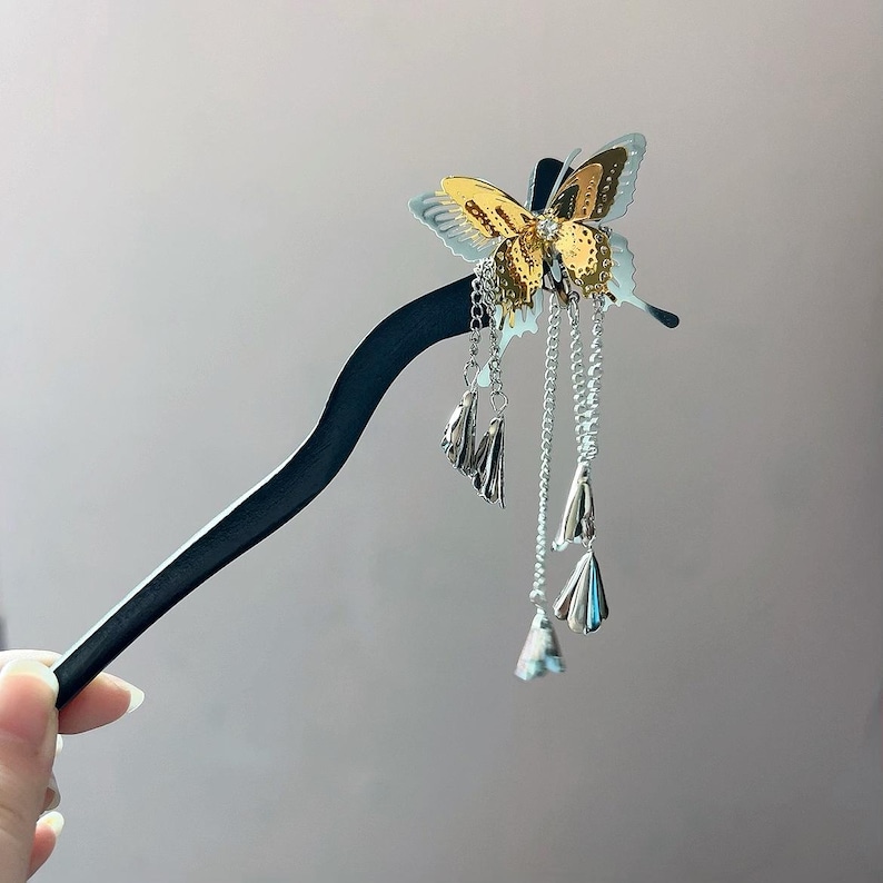 May include: A decorative hair stick featuring a gold and silver butterfly design. The butterfly has intricate details and hangs from a black stick with dangling silver chains and charms. A hand is holding the hair accessory.