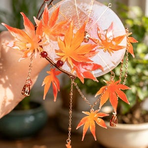 Könnte beinhalten: Ein dekoratives Haaraccessoire mit durchscheinenden orangefarbenen Ahornblatt-Anhängern, einer runden, klaren Harzscheibe und zarten goldfarbenen Metallketten. Das Design umfasst kleine rote Perlen und tropfenförmige Metallanhänger.
