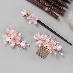 Può includere: Un set di tre accessori per capelli floreali: un bastoncino per capelli, un pettine e una clip. Ogni pezzo presenta delicati fiori rosa con accenti di perle. Il bastoncino per capelli è marrone scuro e il pettine ha denti color oro.