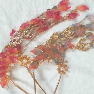 May include: Two ornate hair accessories with leaf-shaped accents in shades of red, orange, and blue. The hairpins feature gold-colored metalwork, delicate chains, and faux pearls. The design is intricate and elegant.