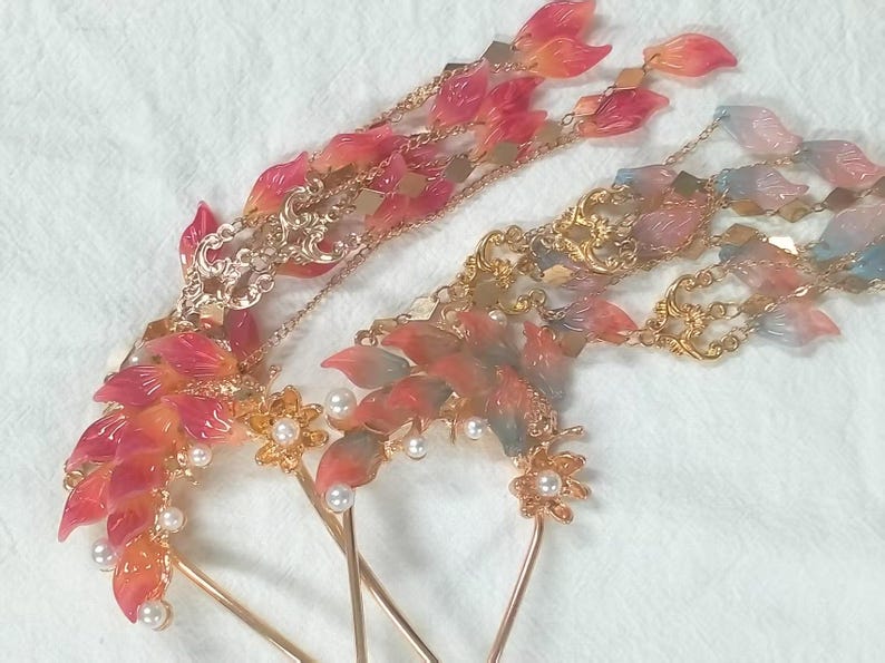 May include: Two ornate hair accessories with leaf-shaped accents in shades of red, orange, and blue. The hairpins feature gold-colored metalwork, delicate chains, and faux pearls. The design is intricate and elegant.