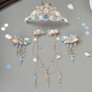 Op de afbeelding: Een verzameling haaraccessoires met zeethema, waaronder een tiara, haarclips, een ketting en oorbellen. De stukken hebben schelpen, parels en blauwe en witte accenten. De tiara staat bovenaan, met de andere stukken eronder.
