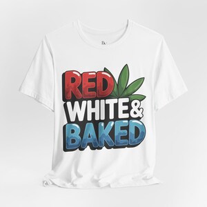 Op de afbeelding: Wit T-shirt met de tekst "RED WHITE & BAKED" in rode, witte en blauwe bubbelletters, met een groene cannabisblad-afbeelding. Het shirt is gemaakt van een zacht materiaal en heeft korte mouwen.