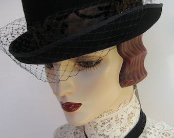 Puede incluir: Un sombrero negro con velo y detalles de plumas. El sombrero tiene una copa redondeada y un ala pequeña. Un velo de red negro cubre la cara. El sombrero está adornado con una pluma y es parte de una colección de moda vintage.