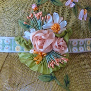 May include: A straw hat with a decorative floral arrangement. The hat features a light green band with a daisy pattern and is adorned with pink and white ribbon flowers, green leaves, and orange accents.