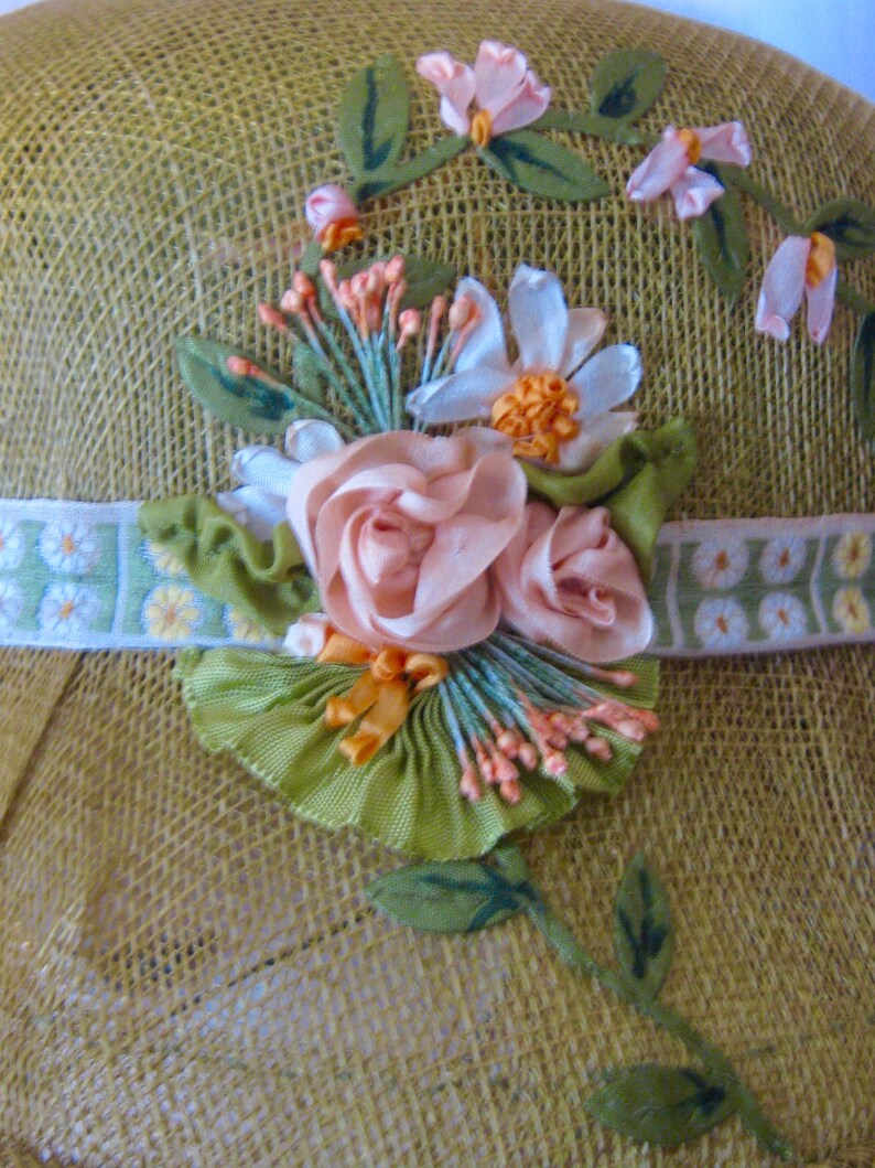 May include: A straw hat with a decorative floral arrangement. The hat features a light green band with a daisy pattern and is adorned with pink and white ribbon flowers, green leaves, and orange accents.