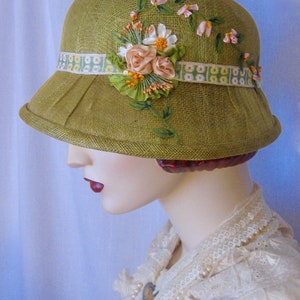 May include: A green cloche hat with a decorative floral arrangement. The hat features a wide brim and a ribbon band with a floral pattern. The floral arrangement includes pink and white flowers with green leaves and stems.