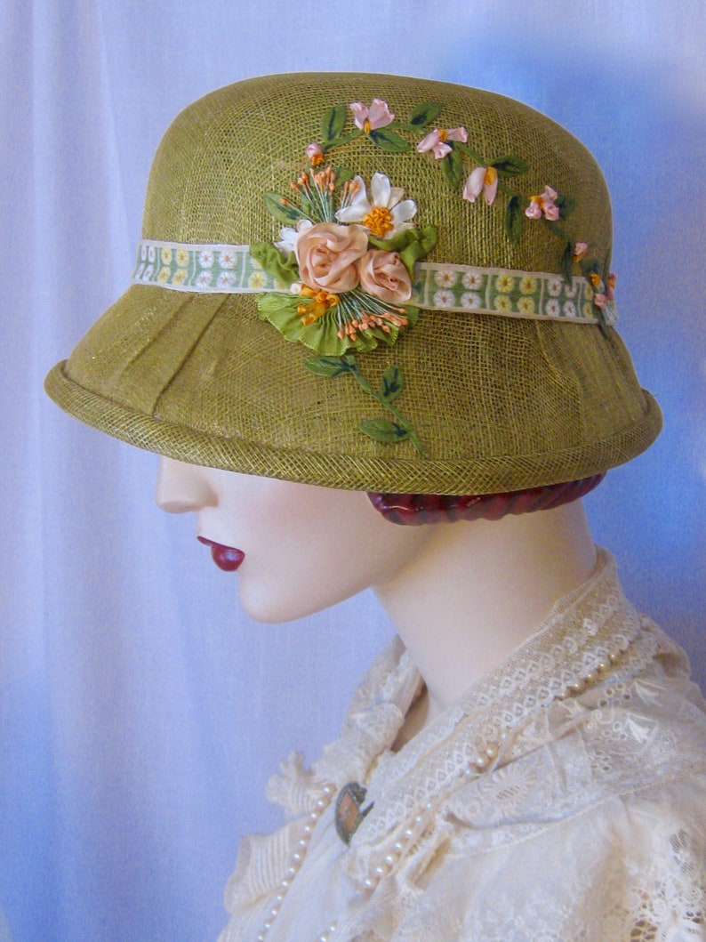 May include: A green cloche hat with a decorative floral arrangement. The hat features a wide brim and a ribbon band with a floral pattern. The floral arrangement includes pink and white flowers with green leaves and stems.