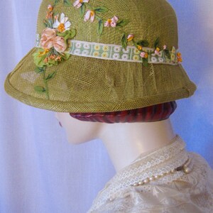 May include: A vintage-style olive green cloche hat. The hat is adorned with a floral ribbon and delicate fabric flowers in shades of pink, white, and yellow. The brim is slightly flared, and the hat has a mesh veil detail.
