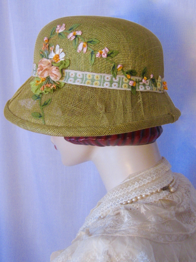 May include: A vintage-style olive green cloche hat. The hat is adorned with a floral ribbon and delicate fabric flowers in shades of pink, white, and yellow. The brim is slightly flared, and the hat has a mesh veil detail.