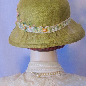 May include: A light green cloche hat with a floral ribbon band. The hat has a pleated brim and is made of a woven material. The ribbon is white with a green and yellow floral pattern and small pink flowers.
