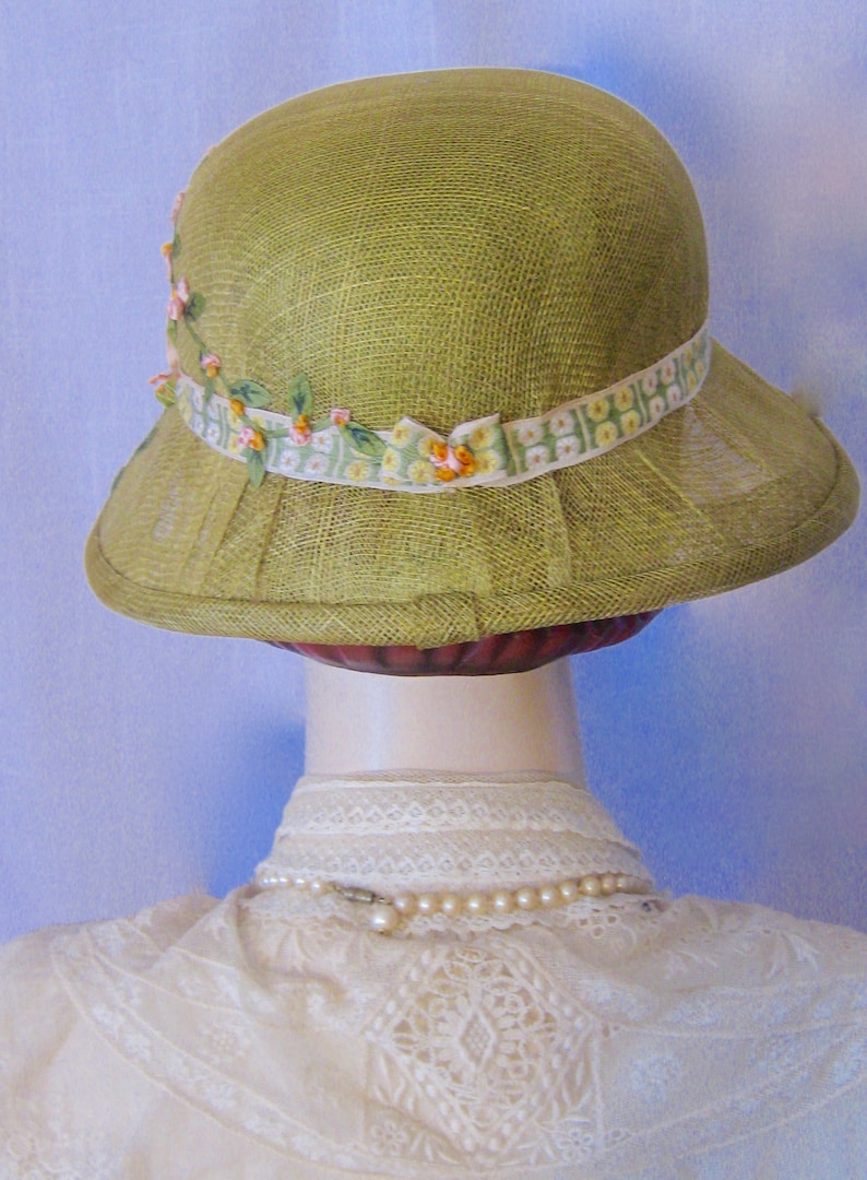 May include: A light green cloche hat with a floral ribbon band. The hat has a pleated brim and is made of a woven material. The ribbon is white with a green and yellow floral pattern and small pink flowers.