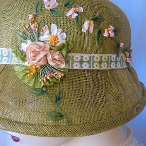 May include: Olive green cloche hat with floral embellishments. The hat features a woven texture and is adorned with pink and white fabric flowers, green leaves, and a decorative ribbon band. A vintage-inspired accessory.