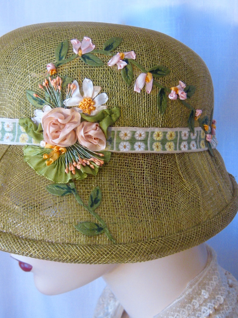 May include: Olive green cloche hat with floral embellishments. The hat features a woven texture and is adorned with pink and white fabric flowers, green leaves, and a decorative ribbon band. A vintage-inspired accessory.