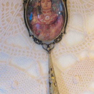 Puede incluir: Colgante de estilo antiguo con un retrato ovalado de una mujer con un atuendo rojo y dorado. El retrato está enmarcado en un metal color bronce con un asa de filigrana decorativa y una gota de cristal azul. El colgante se muestra sobre un fondo de encaje blanco.