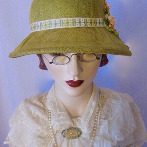May include: A vintage-style, olive green hat with a wide brim and a decorative floral ribbon. The hat is adorned with a cluster of peach-colored flowers. The mannequin is wearing a white lace blouse, a pearl necklace, and a decorative brooch.
