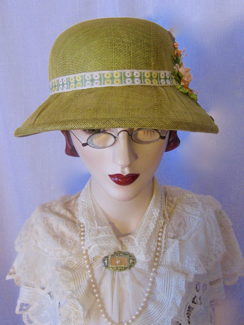 May include: A vintage-style, olive green hat with a wide brim and a decorative floral ribbon. The hat is adorned with a cluster of peach-colored flowers. The mannequin is wearing a white lace blouse, a pearl necklace, and a decorative brooch.