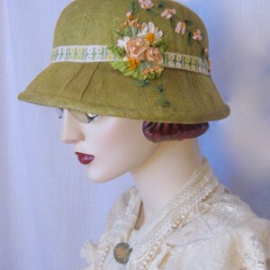 May include: A vintage-style olive green cloche hat with a decorative floral arrangement. The hat features a ribbon band with a floral pattern and is adorned with fabric flowers in shades of pink, peach, and white. The hat has a small brim.