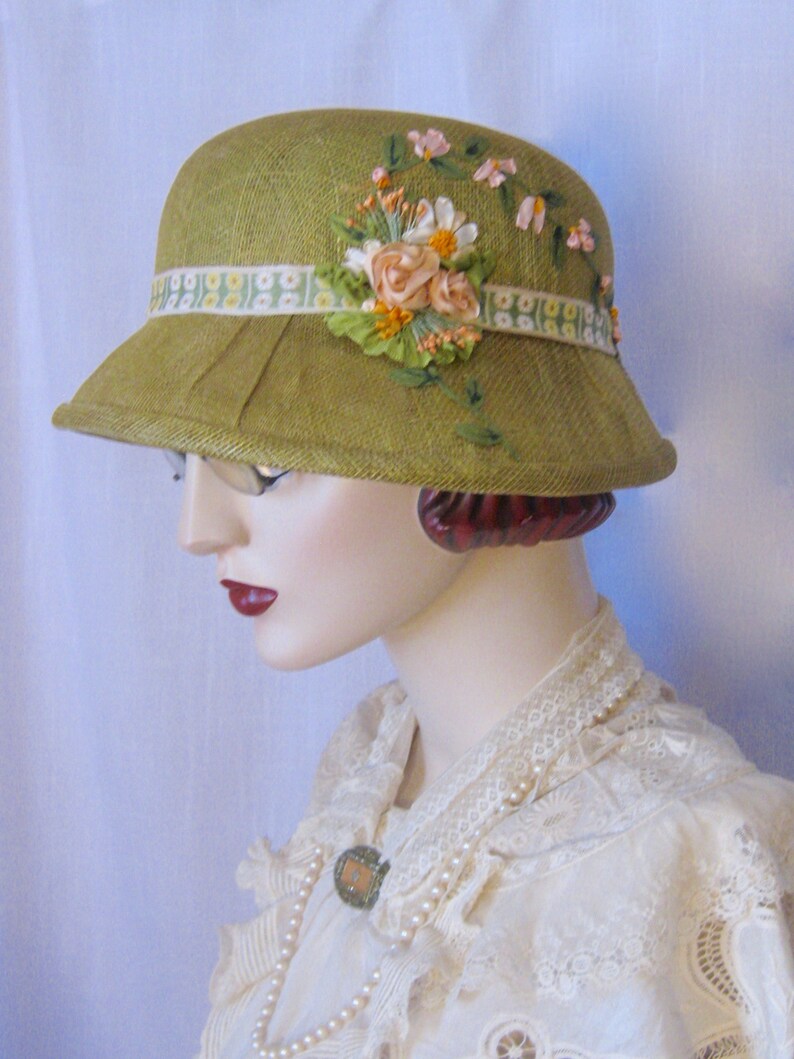 May include: A vintage-style olive green cloche hat with a decorative floral arrangement. The hat features a ribbon band with a floral pattern and is adorned with fabric flowers in shades of pink, peach, and white. The hat has a small brim.