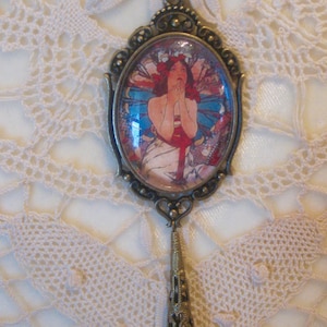 Puede incluir: Un collar con colgante ovalado con un marco de color bronce. El colgante presenta una imagen colorida de una mujer con cabello rojo y un vestido blanco. El collar tiene un cordón negro y una gota decorativa de color bronce.