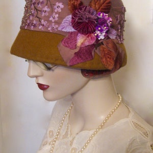 May include: A brown felt cloche hat with a wide brim and a decorative floral embellishment. The embellishment features purple, pink, and brown velvet flowers and leaves.