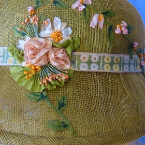 May include: A vintage olive green cloche hat with floral embellishments. The hat features a woven texture and is adorned with pink and white fabric flowers, green leaves, and a ribbon with a daisy pattern. The hat has a rounded crown and a small brim.