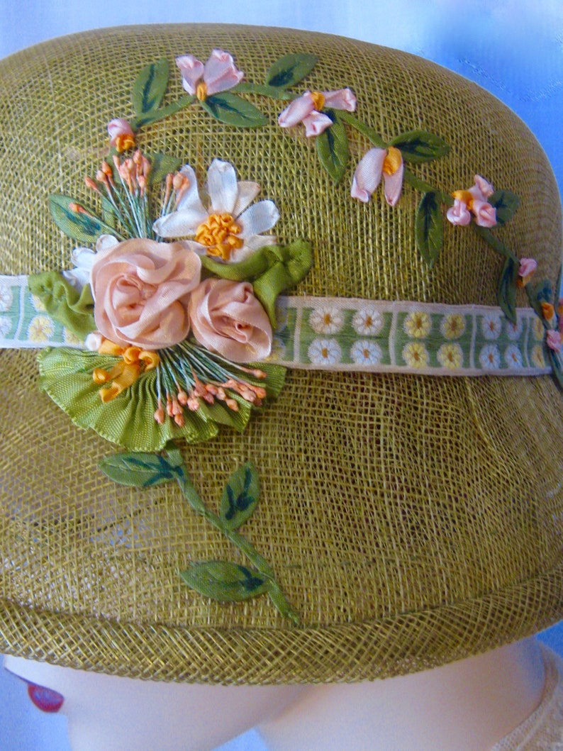 May include: A vintage olive green cloche hat with floral embellishments. The hat features a woven texture and is adorned with pink and white fabric flowers, green leaves, and a ribbon with a daisy pattern. The hat has a rounded crown and a small brim.