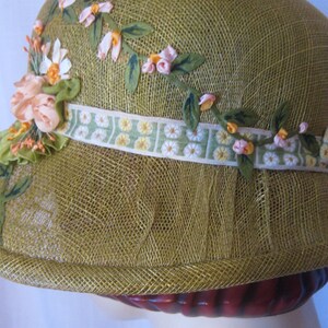 May include: A vintage-style olive green cloche hat. The hat is adorned with floral embellishments, including pink and white fabric flowers, green leaves, and a decorative ribbon with a daisy pattern. The hat is made of a woven material.