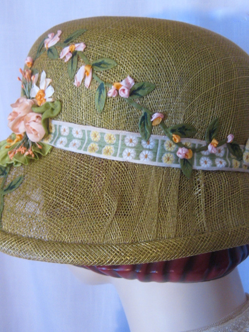 May include: A vintage-style olive green cloche hat. The hat is adorned with floral embellishments, including pink and white fabric flowers, green leaves, and a decorative ribbon with a daisy pattern. The hat is made of a woven material.