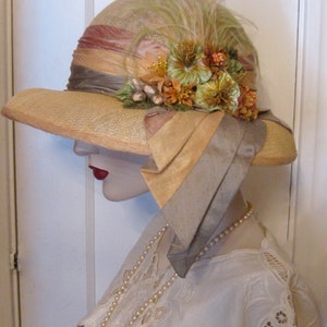 May include: A vintage straw hat with a wide brim, decorated with a band of pink, peach, and brown fabric. The hat is embellished with orange and green flowers, and a feather. A long pearl necklace and white lace top are also visible.