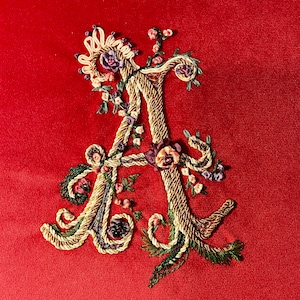 May include: An embroidered letter A in gold and green with pink and purple flowers on a red background.