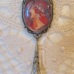 Puede incluir: Colgante de estilo antiguo con una imagen ovalada de una mujer de perfil, en tonos rojos y naranjas. El colgante tiene un marco de metal ornamentado y un largo elemento colgante que termina en una perla nacarada.