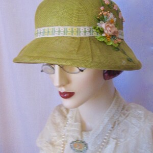 May include: A vintage-style, lime green cloche hat with a decorative floral arrangement and a patterned ribbon band. The hat features a slightly downward-sloping brim and is displayed on a mannequin head. The floral arrangement includes pink and peach-colored flowers.