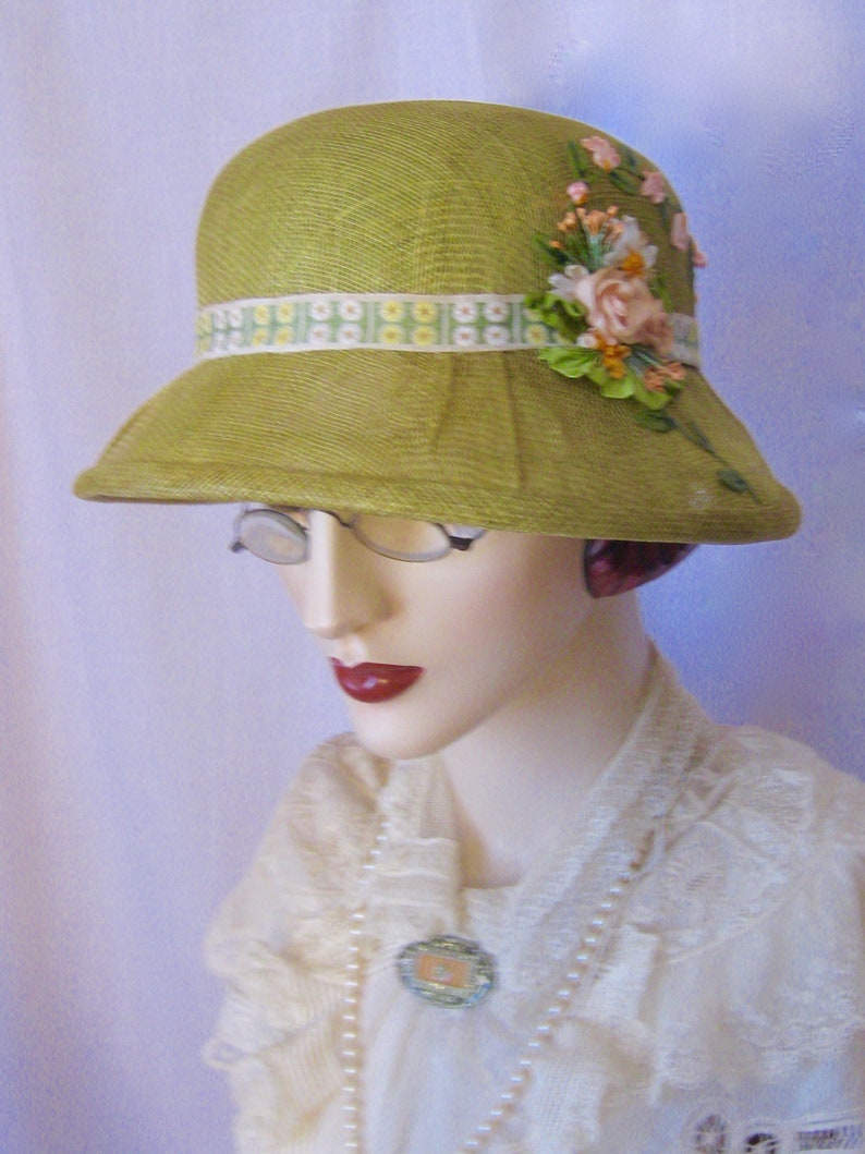 May include: A vintage-style, lime green cloche hat with a decorative floral arrangement and a patterned ribbon band. The hat features a slightly downward-sloping brim and is displayed on a mannequin head. The floral arrangement includes pink and peach-colored flowers.