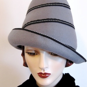 May include: A gray felt hat with a unique shape, featuring black trim with white stitching. The hat has a slightly upturned brim and is worn by a mannequin. The hat's design is accentuated by horizontal bands of trim.