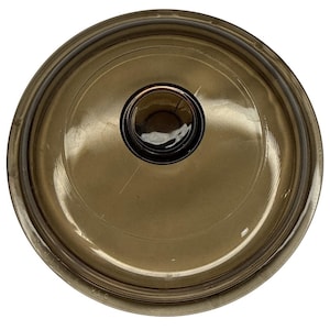 May include: A circular, translucent brown glass lid featuring a smooth, slightly domed surface and a centred, raised knob handle. The lid has a distinct, reinforced outer rim and a polished finish that reflects light across its curved surface.