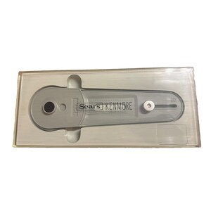 Puede incluir: Un accesorio de máquina de coser de plástico gris claro para ojales, encerrado en un blíster de plástico transparente. La herramienta presenta el texto Sears KENMORE impreso en negro en el centro, con una abertura circular negra y un pasador metálico.