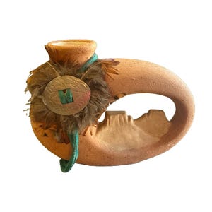 May include: A terracotta-colored ceramic vessel with a curved, hollow handle design. The exterior features a textured surface, a cluster of brown feathers, a circular metal medallion, and a thin teal cord wrapped around the neck.