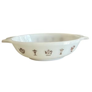 Puede incluir: Una fuente de horno ovalada de vidrio lechoso blanco con dos asas laterales. El exterior presenta un patrón repetitivo de ilustraciones marrones que representan varios utensilios de cocina, incluyendo teteras, cafeteras y cuencos con pie.