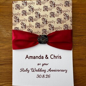 Pode incluir: Um cartão retangular com uma secção superior creme com um padrão castanho repetitivo "Happily Ever After", um laço de cetim vermelho escuro com um broche decorativo escuro e uma secção inferior branca com o texto "Amanda & Chris on your Ruby Wedding Anniversary 30.8.26".