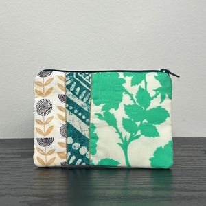 Peut inclure: Une pochette rectangulaire à fermeture éclair et patchwork. La pochette présente des panneaux de tissu avec divers motifs : un motif floral, des formes géométriques turquoise et une impression florale verte. La fermeture éclair est noire.