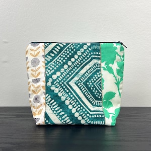 Peut inclure: Une trousse de maquillage patchwork avec une fermeture éclair noire. Le sac présente trois panneaux de tissu avec différents motifs : floral, géométrique et botanique. Les couleurs incluent le blanc, le sarcelle, le vert, l'or et le noir.