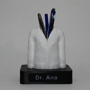Puede incluir: Figurilla de médico de plástico blanco con una base negra. La base tiene el texto "Dr. Ana". La figurita tiene bolígrafos que sobresalen de la parte superior.