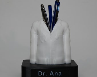 May include: White plastic doctor figurine with a black base. The base has the text "Dr. Ana" on it. The figurine has pens sticking out of the top.