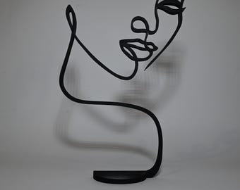 May include: Black metal sculpture of a face in a single line design. The sculpture is on a black base.